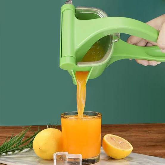 Green manual juicer squeezing orange juice into a glass with lemons on a wooden surface.