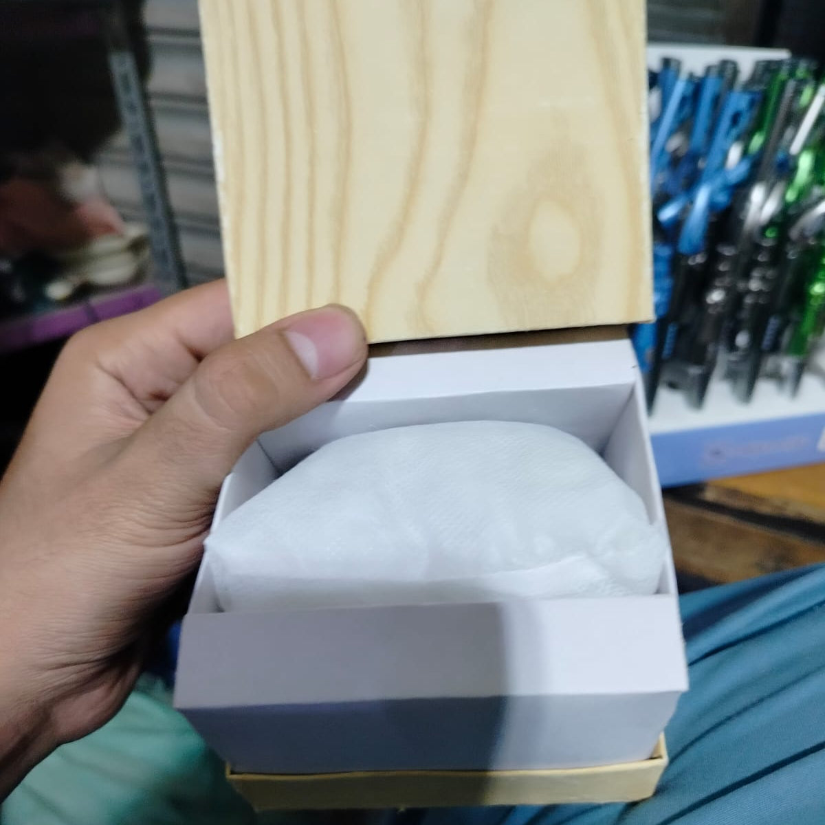 Hand holding a white object in an open box with a wooden block above it, against a blurred background of stationery items.